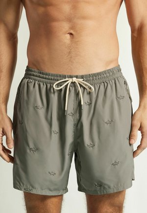 Mann trägt graue Badeshorts mit kleinen schwarzen Hai-Prints und weißem Kordelzug, Oberkörper und Oberschenkel sichtbar.