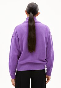 Person mit langen, dunklen Haaren im Pferdeschwanz, der einen lila Rollkragenpullover und schwarze Hosen trägt, von hinten auf einem weißen Hintergrund gezeigt.