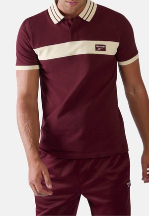 Mann trägt ein burgunderfarbenes Polohemd mit beigem Streifen und Kragen, kombiniert mit passenden burgunderfarbenen Hosen, beide mit Reebok-Logos.