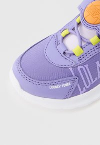 Zapatilla deportiva morada con parte superior de tela, con el logo de "LOONEY TUNES", detalles en amarillo y suela de goma blanca. Primer plano de la zona de la puntera.
