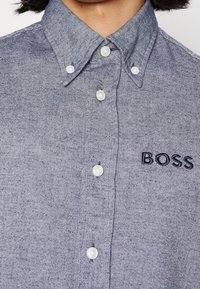 Košile na knoflíky z modrého chambray materiálu s bílými knoflíky. Na levé straně hrudi se nachází vyšívané logo "BOSS" v tmavě modré barvě.