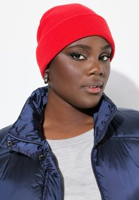 Berretto a coste rosso indossato sopra una maglietta grigia e un giubbotto puffato blu navy. Il giubbotto presenta una chiusura con zip e bottoni a scatto con una texture lucida.