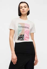 Νεαρή γυναίκα που φοράει λευκό T-shirt με πολύχρωμο ζωικό μοτίβο και τη λέξη «Εμπιστοσύνη», συνδυασμένο με μαύρη φούστα με ψηλή μέση και σκίσιμο.