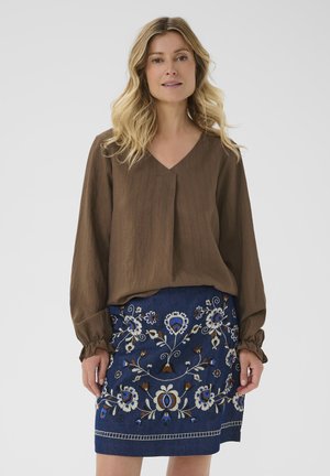Femme avec des cheveux ondulés blonds portant un blouse à manches longues marron et une jupe bleue ornée de broderies florales multicolores, se tenant devant un fond blanc.