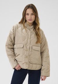 Quilted beige jacket with a high collar, snap-button front, and two front pockets; smooth texture and relaxed fit.
