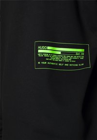 T-shirt in cotone nero con etichetta stampata fluorescente verde contenente testo e elementi di design, centrata nell'area sinistra del petto.