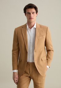 Completo di lino color beige chiaro con rever a punta, chiusura a un bottone e due tasche frontali. Abbinato a una camicia bianca con bottoni.