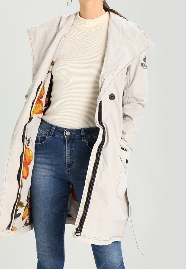 Giacca da esterno grigia chiara con cappuccio, con fodera floreale a fantasia, chiusura con bottoni e tasche laterali. Indossata sopra un maglione beige.