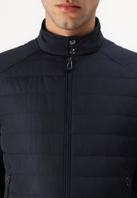 Giacca trapuntata blu navy con colletto alto, chiusura con zip e bottoni a pressione. Presenta pannelli a contrasto sulle spalle e una texture liscia.