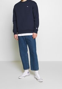 Sudadera navy con cuello barco, puños de canalé y pequeño logo. Combinada con jeans azules holgados y zapatillas deportivas blancas de caña alta.