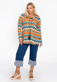 Multicolore tuniek met zigzagpatroon in oranje, teal en zwart; lange mouwen; gecombineerd met cropped denim broeken met een omgeslagen zoom en wedges.