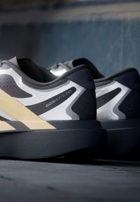 Ζευγάρι ασπρόμαυρα παπούτσια για τρέξιμο Adidas Adizero Evo SL Exo τοποθετημένα παράλληλα σε σκοτεινή επιφάνεια.