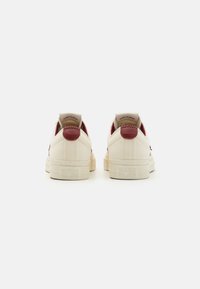 Beige canvas sneakers with maroon heel and logo accents. Low-top design with textured canvas, subtle stitching, and rubber outsole.