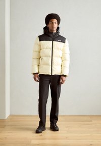 Giacca gonfiabile di colore giallo chiaro con cappuccio e spalle nere; presenta chiusura con zip, tasche laterali e una texture trapuntata. Abbinata a pantaloni cargo neri e scarpe.