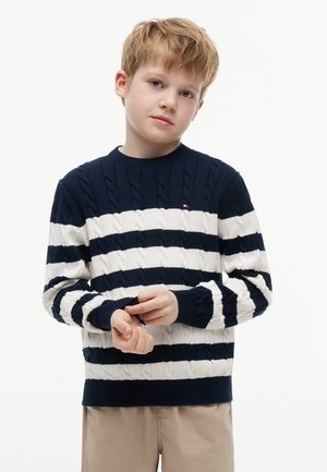 Lille dreng med lysebrunt hår, iført en marineblå og hvidstribet sweater og beige bukser, justerer sin ærme mod en ensfarvet baggrund.