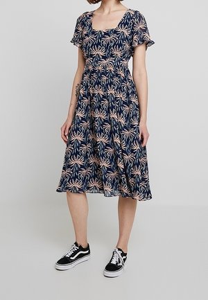 Floral print dress featuring a square neckline, short flutter sleeves, and a cinched waist, in navy blue with pink leaves. Paired with black sneakers.