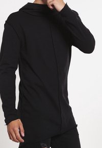 Camiseta negra de manga larga con capucha, hecha de un tejido suave. Presenta un dobladillo asimétrico único y un detalle de costura en la parte delantera.