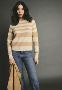 Pull rayé beige et crème avec un ourlet côtelé, associé à un jean bleu à jambes larges. Une veste en daim beige est tenue dans la main du mannequin.