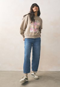 Jasnobrązowa bluza z nadrukiem graficznym ust; luźny krój. Niebieskie dżinsy o prostych nogawkach; kremowo-czarne tenisówki na sznurówki.