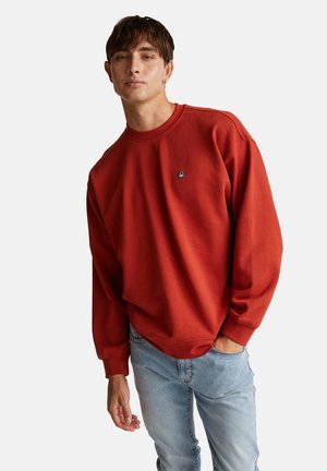 Rode sweatshirt met een ronde hals, geribbelde manchetten en een klein geborduurd logo op de borst. Gecombineerd met lichtblauwe spijkerbroek. Zachte textuur.
