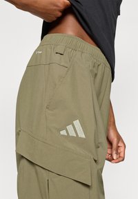 Χακί ελαστικό παντελόνι cargo με ελαστική μέση, πλαϊνές τσέπες και ανακλαστικό λογότυπο Adidas. Κατασκευασμένο από ελαφρύ υλικό με λεία υφή.