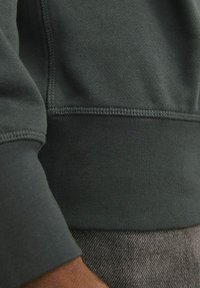 Donkergroene sweatshirt van een zachte, textuurrijke stof. Bevat ribgebreide manchetten en zoom met zichtbare stikseldetails. Close-up weergave van de arm.