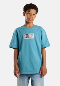 Tiffanysin värinen lyhythihainen puuvillapaita, jossa on suorakulmainen tarra, jossa on numero 89 ja teksti "New York". Suora malli ja pyöreä pääntie.