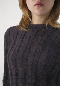 Frau, die einen dunkelgrauen, kuschelig strukturierten Pullover mit Zopfmuster trägt, ihr Mund und unteres Gesicht vor einem einfarbigen Hintergrund zeigend.