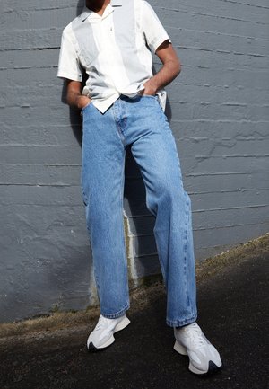 Person wearing light blue wide-leg jeans, white sneakers, and a short-sleeve collared shirt standing against a textured gray wall.