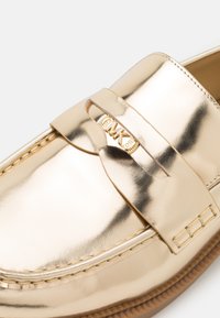 Gouden leren loafer met een decoratieve band met een gouden logo, een gladde afwerking, gestikte details langs de randen en een rubberen zool.