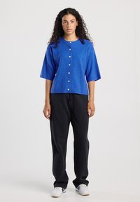 Vrouw met krullend haar in een blauwe blouse met knopen en korte mouwen, zwarte broek en witte sneakers, staande tegen een effen lichte achtergrond.
