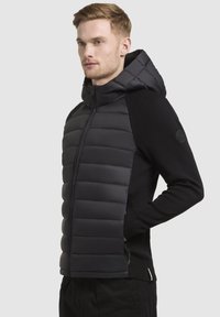 Gilet imbottito nero con cappuccio, maniche a maglia a contrasto e chiusura con zip. Presenta una finitura testurizzata e il logo sulla manica.