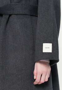 Grå uldfrakke med bælte, med et firkantet Calvin Klein mærke på ærmet. Blød tekstur, brede ærmer og en skræddersyet design.