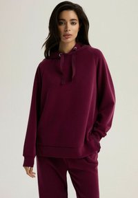 Bordowa bluza z kapturem wykonana z miękkiego materiału, o luźnym kroju, z dekoltem ze ściągaczem i ściągaczami przy rękawach. Zestawiona z pasującymi spodniami.