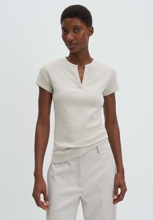 Donna che indossa una maglietta beige chiaro a maniche corte a coste con scollo a spacco e pantaloni beige chiaro abbinati, in piedi contro uno sfondo semplice.