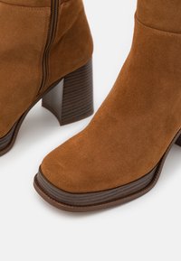 Stivali ankle in suede marrone con punta quadrata, dotati di un tacco in legno texturizzato e chiusura con zip laterale per una calzata facile.