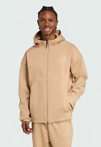 Μπεζ φούτερ με φερμουάρ φτιαγμένο από μαλακό υλικό, με κουκούλα, δύο μπροστινές τσέπες και ένα μεγάλο τονικό λογότυπο στο αριστερό στήθος.