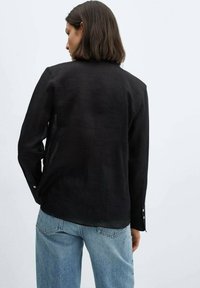 Kvinde med skulderlangt brunt hår iført en sort langærmet skjorte og lyseblå jeans, vendt væk mod en ensfarvet baggrund.