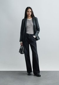 Ženska stoji, oblečena v črno usnjeno jakno, sivo rebrasto majico in črne hlače širokih hlačnic, drži majhno črno torbico, v ozadju je enobarvna siva stena.