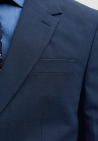 Κοντινό πλάνο της πέτου ενός σκούρου μπλε σακακιού, τσέπης στο στήθος και μέρους ενός γαλάζιου πουκάμισου με ορατή σκούρα γραβάτα με σχέδιο.