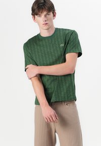 Tricou verde cu mânecă scurtă, având un model geometric, guler rotund și logo brodat. Purtați cu pantaloni din bumbac bej deschis cu nervuri.