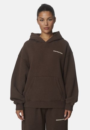 Donna con capelli raccolti che indossa una felpa oversize marrone scuro con cappuccio e pantaloni abbinati con piccoli loghi bianchi di testo sul petto e sulla coscia.