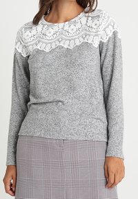 Femme portant un pull gris à manches longues avec de la dentelle florale blanche sur le haut de la poitrine et les épaules, assorti à une jupe à carreaux.