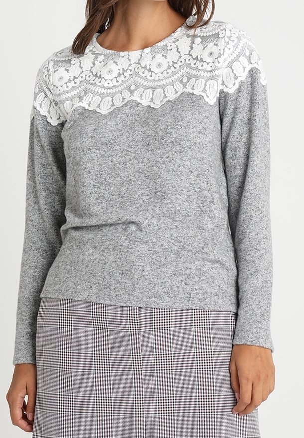 Femme portant un pull gris à manches longues avec de la dentelle florale blanche sur le haut de la poitrine et les épaules, assorti à une jupe à carreaux.