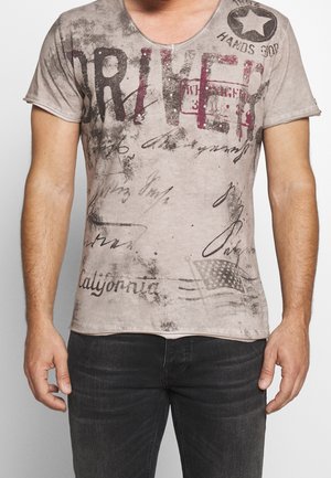 Mann trägt ein kurzärmeliges, ausgewaschenes graues T-Shirt mit "DRIVEN" Text, Stern-Emblem, amerikanischer Flagge und verschiedenen Schriftzügen, kombiniert mit dunklen Jeans.