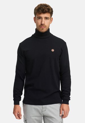 Pull à col roulé noir en tissu tricoté texturé avec un petit logo rond sur la poitrine. Manches longues et design ajusté, associé à un pantalon gris clair.