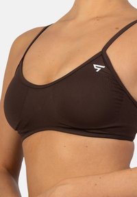 Ruskea saumaton bralette ohutolkaimilla, jossa on valkoinen logo sivussa. Valmistettu pehmeästä, joustavasta materiaalista, jonka pinta on sileä.