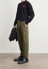 Sweat-shirt noir avec le logo rouge « HUGO », associé à un pantalon cargo vert olive avec poches latérales. Des chaussures en cuir noires complètent la tenue.