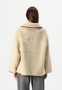 Beige nepleren jas met een brede kraag, lange mouwen en een ontwerp van een achternaad. Bruine piping accentueert de randen. Zachte textuur.