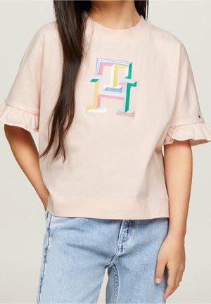 Dziewczyna ubrana w jasnoróżową bluzkę z krótkim rękawem wykończoną falbankami, z kolorowym haftowanym literą "F" na przodzie, w połączeniu z jasnoniebieskimi dżinsami.